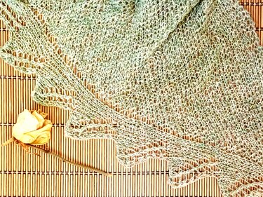 Gestricktes Dreieckstuch mit lockerem Lochmuster und gezackter Bordüre, liegend auf einer Bambusmatte neben einer getrockneten gelben Rose