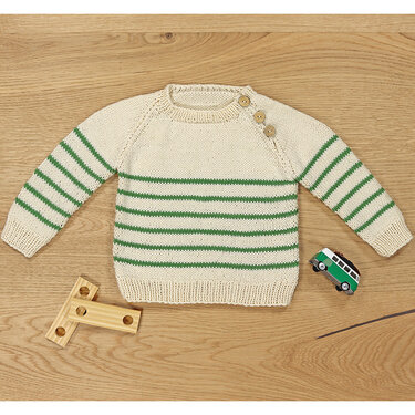 Cremefarbener Kinderpullover mit grünen Streifen und drei Holzknöpfen an der Schulter, auf Holztisch liegend.