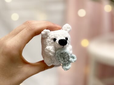 Hand hält kleines schnell gehäkeltes weißes Amigurumi-Bärchen mit einem gehäkelten Deko-Element