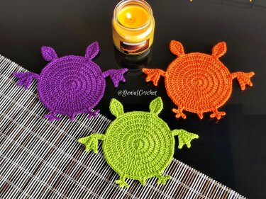 Three round crochet monster coasters in purple, orange and lime on a black table next to a lit candle and placemat.