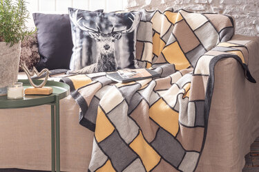 Gehäkelte Patchwork-Decke in Senf, Grau und Beige auf einem hellen Sofa mit Kissen und Beistelltisch