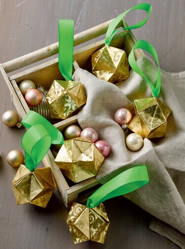 Goldene gefaltete Origami-Anhänger mit grünen Satinbändern in einer Holzbox neben pastellfarbenen Weihnachtskugeln auf Leinen