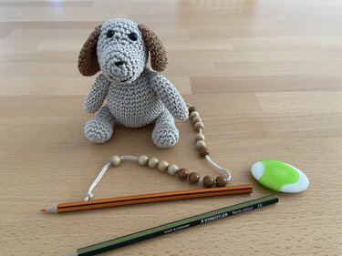 Kleiner gehäkelter Hund mit braunen Schlappohren neben zwei Buntstiften, Radiergummi und Holzperlen auf Holztisch.