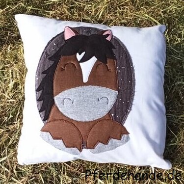 Weißes Kissen mit appliziertem braunem Comic-Pferd auf dunklem, sternenbesetztem Oval, liegend auf Gras