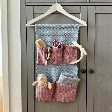 Gehäkelter Organizer mit mehreren Taschen in Rosa und Hellblau an einem Kleiderbügel