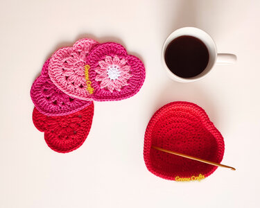 Overhead view of pink and red crocheted heart coasters, a red heart-shaped crochet basket with a hook, and a white coffee mug.