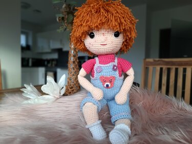 Handgehäkelte Amigurumi-Puppe mit roten Fusselhaaren, Sommersprossen, rosa Shirt und blauer Latzhose mit Bärenapplikation auf rosafarbenem Fell