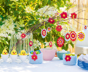Oster-Häkeldeko auf dem Tisch: blaue Häkelkörbchen mit Blüte, Küken-Eierwärmer in Eierbechern und runde Mandala-Anhänger am Zweig