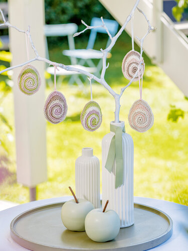 Gehäkelte Ostereier in Pastellfarben in Spiralform, hängend an einem weiß gestrichenen Ast, mit Vasen und Dekoäpfeln auf einem Tablett.