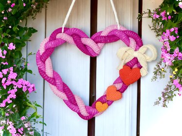 Heart-shaped braided pink and fuchsia crochet wreath with a cream bow and small red and orange crochet hearts, hanging on a white door surrounded by pink flowers.