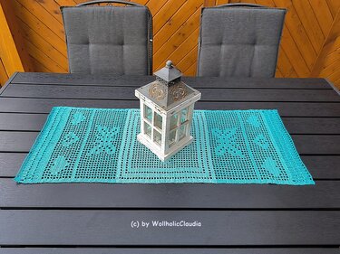 Gehäkelter türkiser Tischläufer mit Filetmuster und weißer Laterne auf dunklem Holztisch.