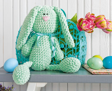 Mintgrüner gehäkelter Amigurumi-Hase aus Plüschgarn mit langen Schlappohren und Schleife neben Ostereiern