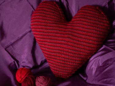 Red striped knitted heart pillow on purple fabric with yarn balls