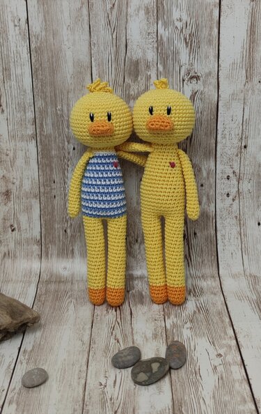 Zwei gelbe gehäkelte Amigurumi-Enten, eine mit blau-weiß gestreiftem Oberteil, beide mit kleinen roten Herzen, vor hellem Holzuntergrund.