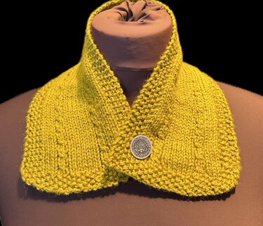 Senfgelber gestrickter Kragen mit dekorativem Knopf auf einer braunen Büste.