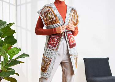 Person trägt lange, ärmellose Häkelweste aus Granny-Quadraten in Hellgrau mit orange-braunen Akzenten über einem Rollkragenpullover.