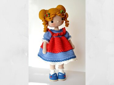 Full-body amigurumi doll with yellow curly hair, blue dress, red pinafore, white socks, and blue buttoned shoes on a light background.