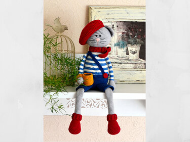 Grey crocheted amigurumi cat wearing a red beret and scarf, striped shirt, blue overalls, red boots, holding an orange mug.