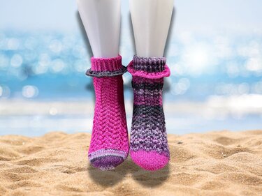 Zwei kurze, gestrickte Stulpen in Pink und Lila an Schaufensterpuppenfüßen auf Sand vor Meereshintergrund.