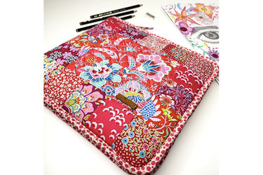 Gesteppte Patchwork-Tasche in Rottönen mit floralen Mustern, liegend neben Bleistiften und einer Zeichnung.