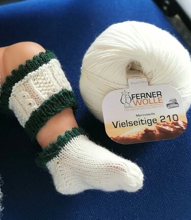 Gestrickte Babysocken und Beinstulpen in Creme mit grünem Rand neben einem Knäuel weißer Wolle