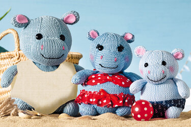Drei gestrickte Nilpferd-Amigurumi in Strand-Outfits sitzen im Sand