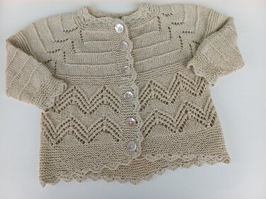 Beige gestrickte Babyjacke mit Zickzack-Lochmuster, Rippenstruktur oben und fünf Perlmuttknöpfen, flach auf hellem Hintergrund.