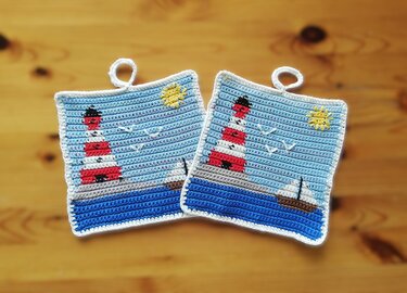 Paar gehäkelter quadratischer Topflappen mit Leuchtturm, Segelboot, Möwen und Sonne vor blauem Meer