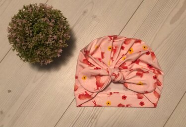 Nahaufnahme einer rosa Knotenmütze mit vorne gebundenem Knoten, Umschlagbund und kleinem Tier- und Blumenmuster auf hellem Holzboden