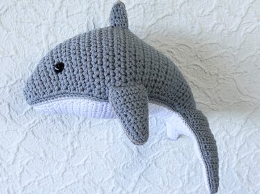 Gehäkelter grauer Amigurumi-Wal mit weißem Bauch vor strukturierter Wand