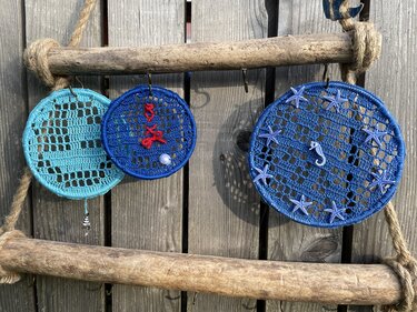 Drei gehäkelte Ringe in Türkis und Blau unterschiedlicher Größe hängen an einer Treibholzstange, dekoriert mit Seestern-, Seepferdchen- und Muschelanhängern