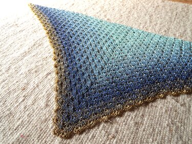 Dreieckiges gehäkeltes Tuch mit blau-türkis Farbverlauf und goldfarbener Muschelkante auf hellem Teppich