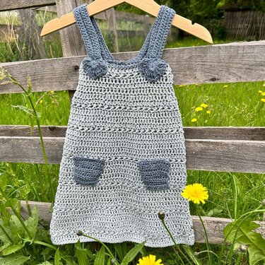 Hellblaues handgehäkeltes Kinderkleid mit dunkleren Trägern und zwei aufgesetzten Taschen, an einem Holzbügel an einem Holzzaun aufgehängt.