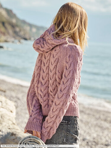 Person von hinten in altrosa Kapuzenpullover mit Zopfmuster am Strand, Meer und Felsen im Hintergrund.