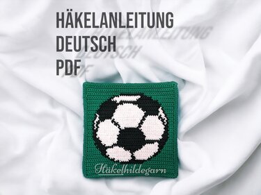 Quadratischer gehäkelter Untersetzer mit schwarz-weißem Fußballmuster auf grünem Hintergrund.