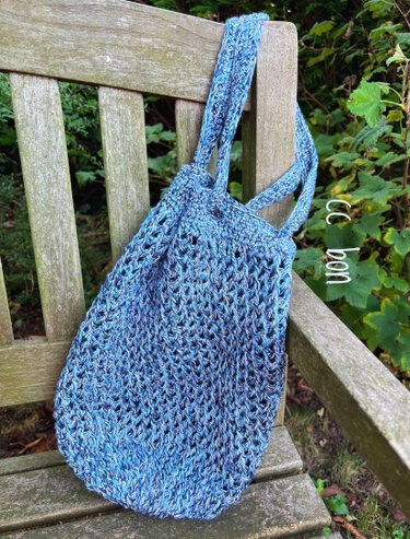 Blauer gehäkelter Shopper mit Lochmuster an Holzbank hängend.