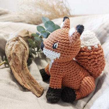 Gehäkelter Fuchs‑Amigurumi, orangebraun mit weißer Schnauze und Schwanzspitze, sitzt auf hellem Stoff.