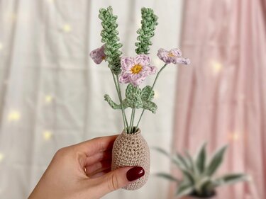 Hand hält kleine beigefarbene gehäkelte Vase mit hellvioletten Blüten und grünen Stielen.