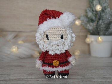 Gehäkelter Weihnachtsmann in roter Kleidung mit weißem Bart vor unscharfem weihnachtlichem Hintergrund