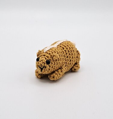 Small brown crocheted amigurumi animal with a white stripe, black safety eyes, and a stitched smile on a white background.