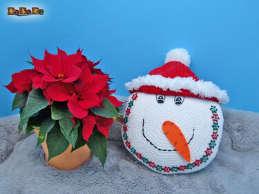 Rundes, gehäkeltes Schneemann-Kissen mit roter Weihnachtsmütze, Karottennase und nummeriertem Sternenrand neben einem roten Weihnachtsstern.