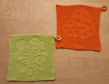 Zwei quadratische gestrickte Topflappen, hellgrün mit Kleeblattmuster und orange mit Baum-Motiv, jeweils mit Aufhängeschlaufe, auf hellem Holzuntergrund