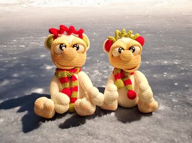 Zwei handgehäkelte Amigurumi mit rot-grün gestreiften Schals auf frostigem Untergrund.