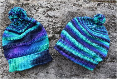 Zwei gestrickte Kindermützen mit Bommel in Blau-, Türkis- und Lilatönen auf grauem Steinuntergrund
