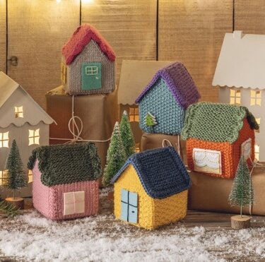 Gestrickte Minihäuser in verschiedenen Farben, arrangiert auf Geschenken und Schnee mit kleinen Tannen.