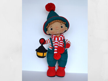Amigurumi boy doll in teal hat with red pom-pom, striped red-and-white scarf, teal overalls and red boots, holding a small yellow-and-black lantern.