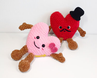 Two whimsical crochet heart amigurumi, pink with bow and smile and red with mustache and top hat, sitting side by side