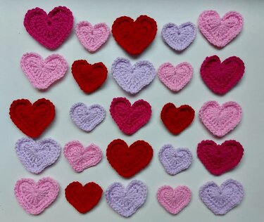 25 crocheted heart appliqués in red, pink and lavender arranged on a white background