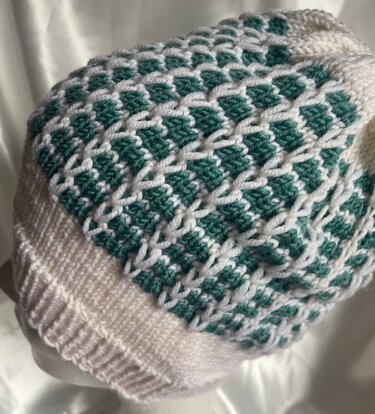 Nahaufnahme einer gestrickten Mütze in Weiß und Dunkelgrün mit geometrischem Wabenmuster und geripptem Bund