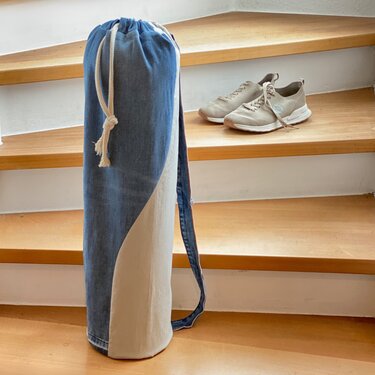 Hohe, schmale Zylindertasche aus blauem Jeansstoff und beigem Stoff mit Kordelzug und Schultergurt auf einer Holztreppe neben beigen Sportschuhen.